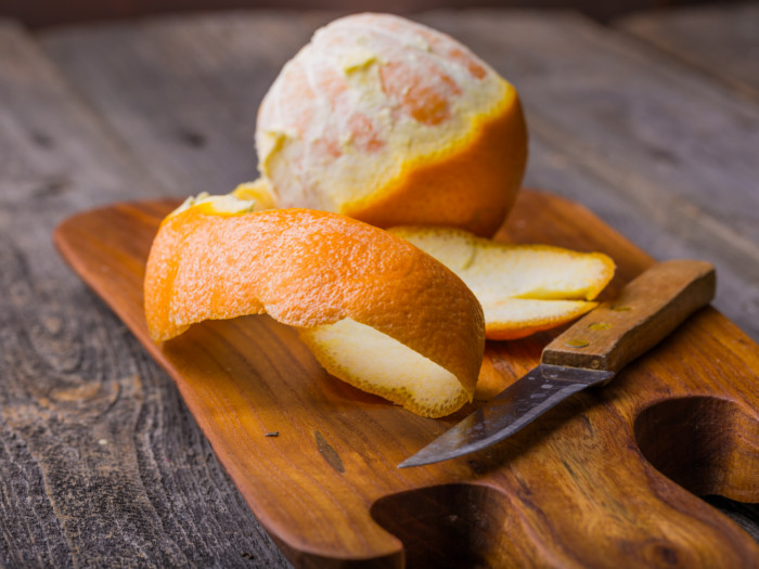 An orange peel, a knife and a partially peeled orange on a wooden board on a wooden surface